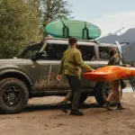 Ford Bronco am Bergsee während zwei Personen ein Kajak und Paddel tragen