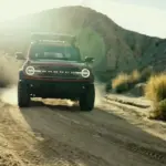 Ford Bronco fährt frontal auf einer staubigen Wüstenpiste zwischen Felsen